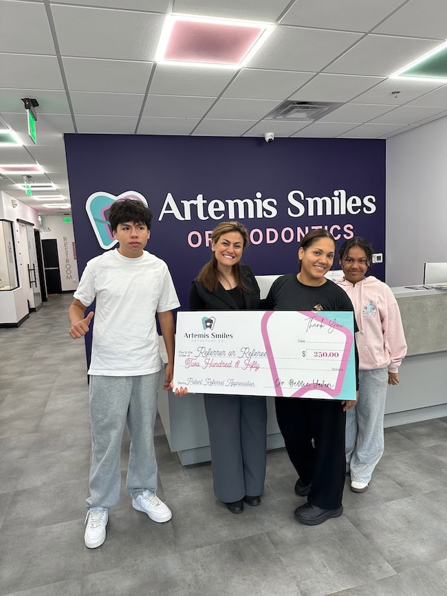 Smiling teens holding braces-off certificates in front of the Artemis Smiles Orthodontics sign
